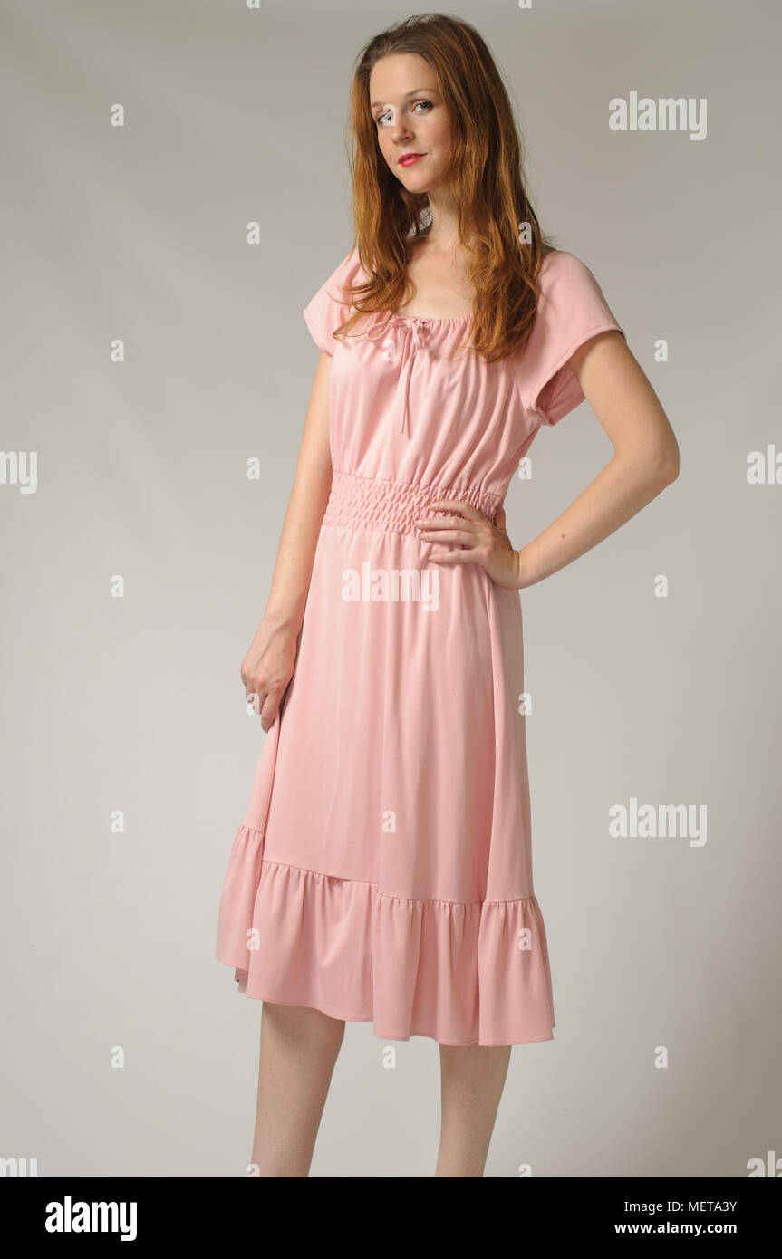 Red hair female modeling posing in front of a grey background wearing a pink vintage dress. A vintage fashion portrait concept. Stock Photo