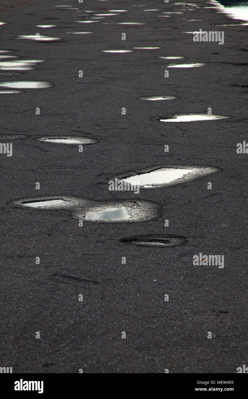 A cloudy sky is reflected in rain puddles that have gathered in holes in the asphalt covering a road. Stock Photo