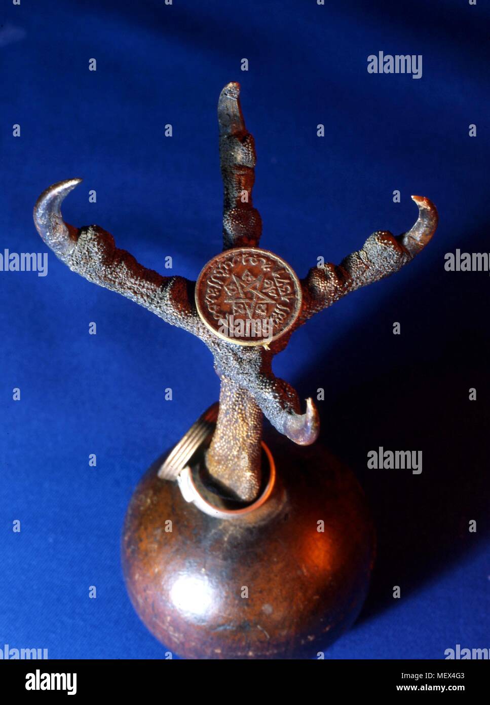Open claw of bird of prey, mounted on wooden ball base with Arabic coin (Hijri date 1371, 1951 in Gregorian calendar), bearing pentagram, stuck to centre of claw. 17 cms in height. c. 1950s, or older in part. Thought to have been used in the process of invoking djinn by bating them into a trap. Stock Photo