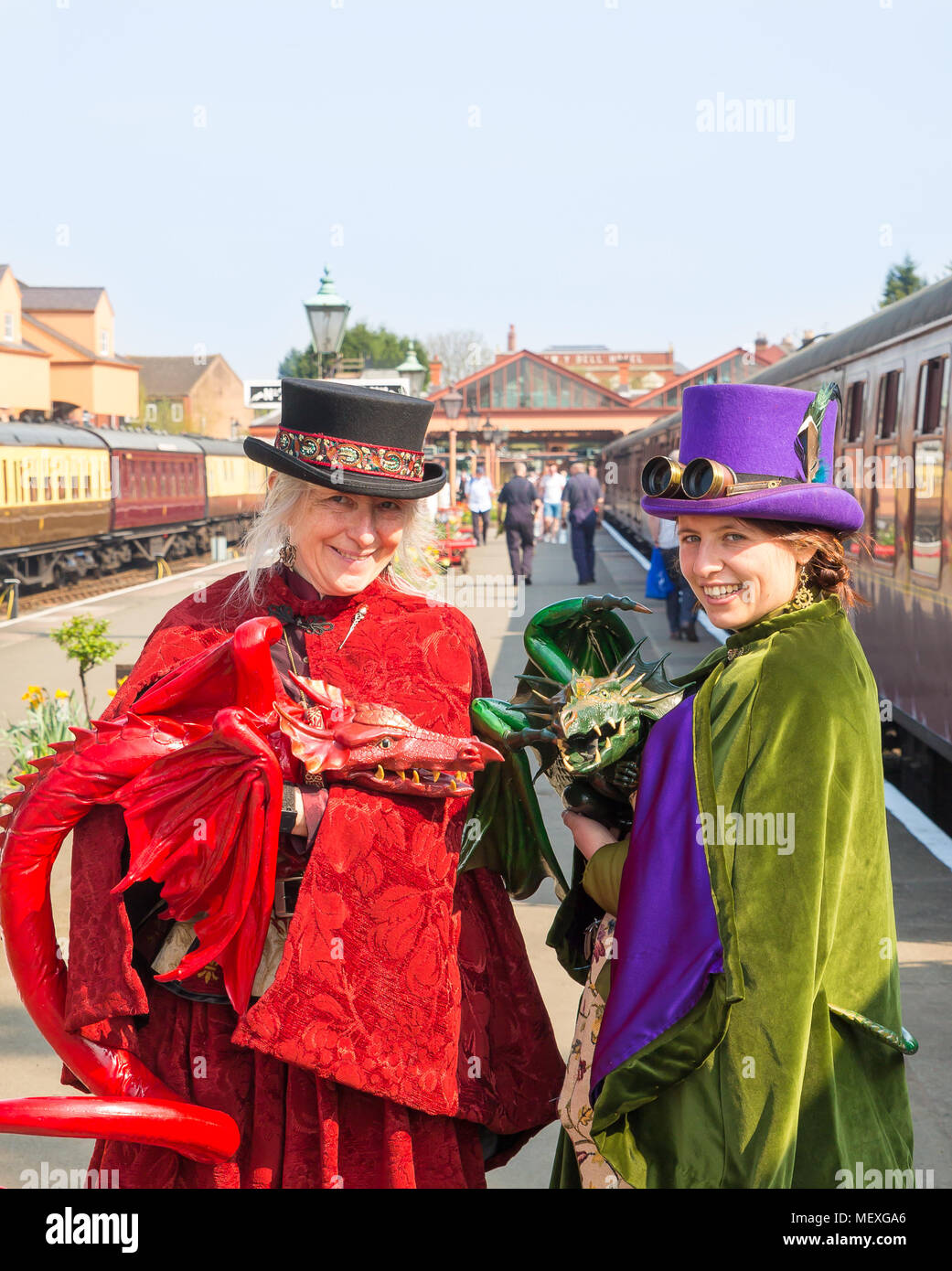 Portrait shot of two smiling females in fancy dress costume with hand-held, life-like dragon puppets on platform in sun, at SVR Kidderminster station. Stock Photo