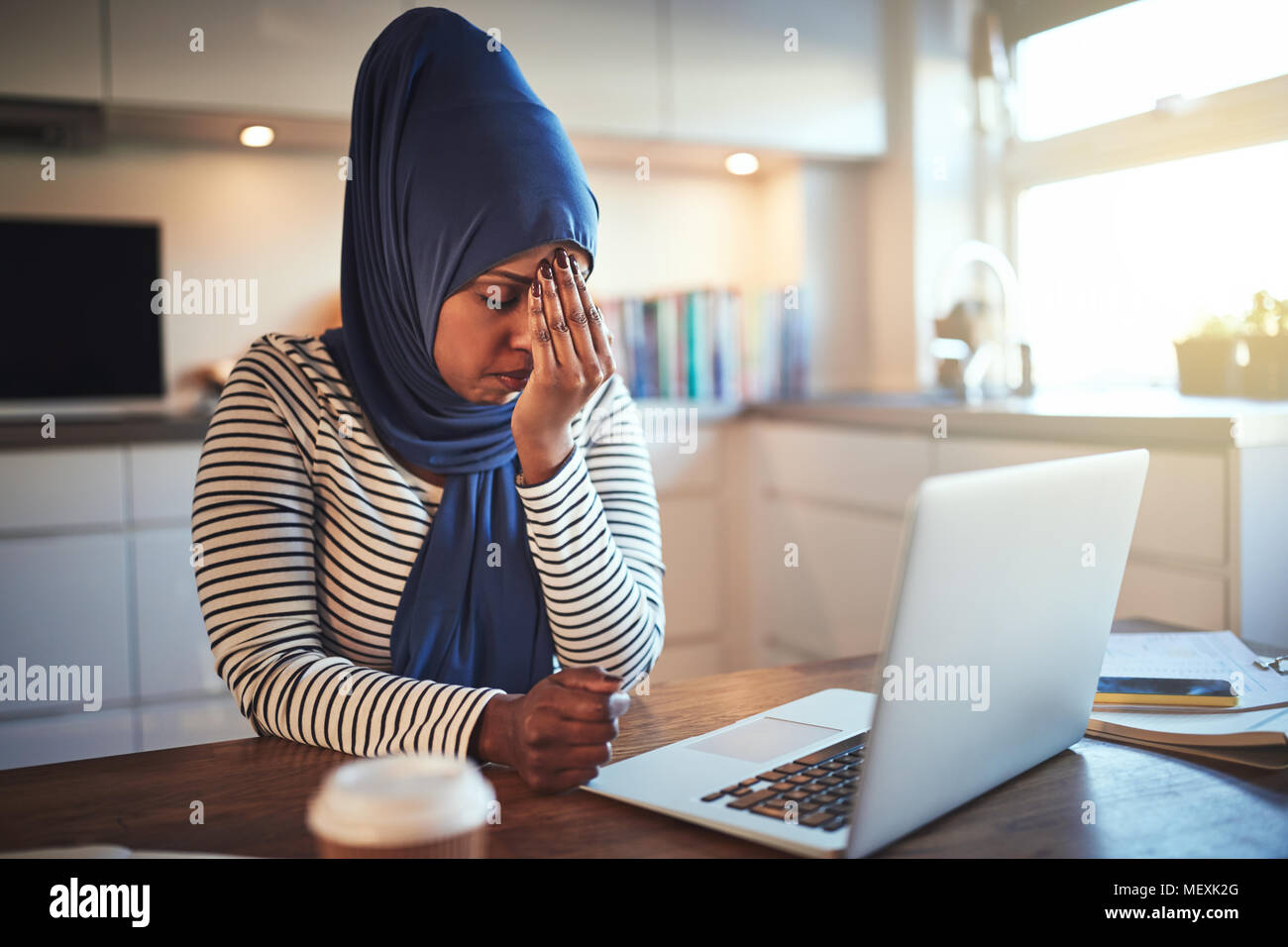 Stressed out young Arabic female entrepreneur wearing a hijab sitting at her kitchen table working online with a laptop Stock Photo