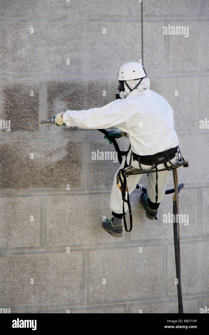 Rope access facade maintenance; A worker wearing a protective gear cleaning a stone church exterior with abrasive blasting equipment Stock Photo