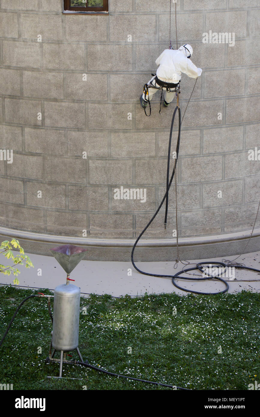 Rope access facase maintenance; A worker wearing a protective gear cleaning a stone church exterior with abrasive blasting equipment Stock Photo