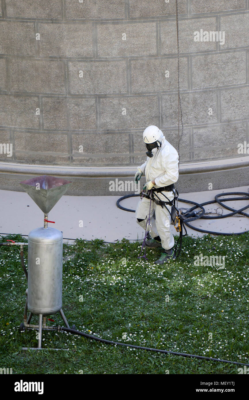 Rope access facase maintenance; A worker wearing a protective gear cleaning a stone church exterior with abrasive blasting equipment Stock Photo