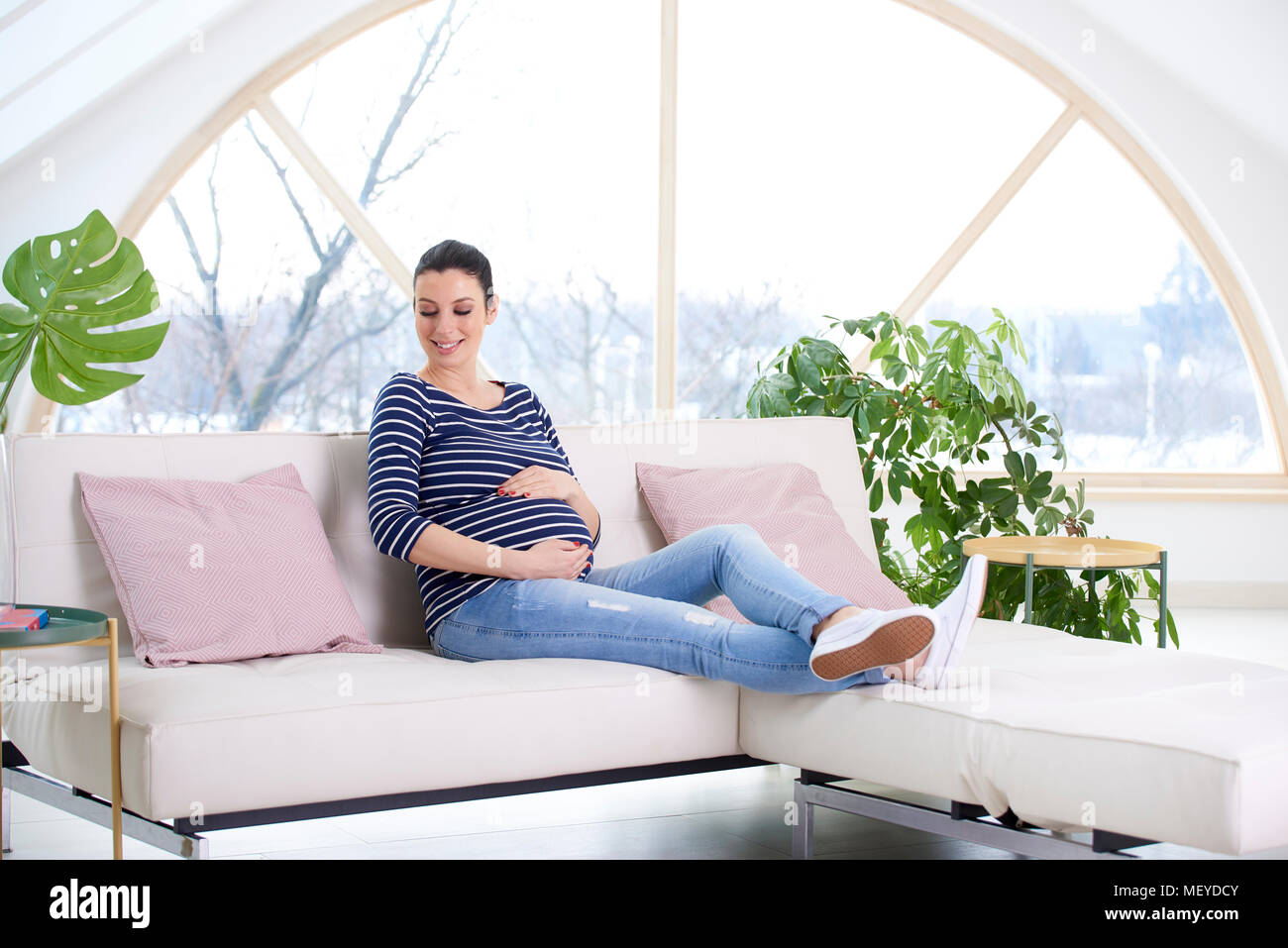Full length shot of beautiful pregnant woman sitting on sofa and touching  her baby bump while relaxing at home Stock Photo - Alamy