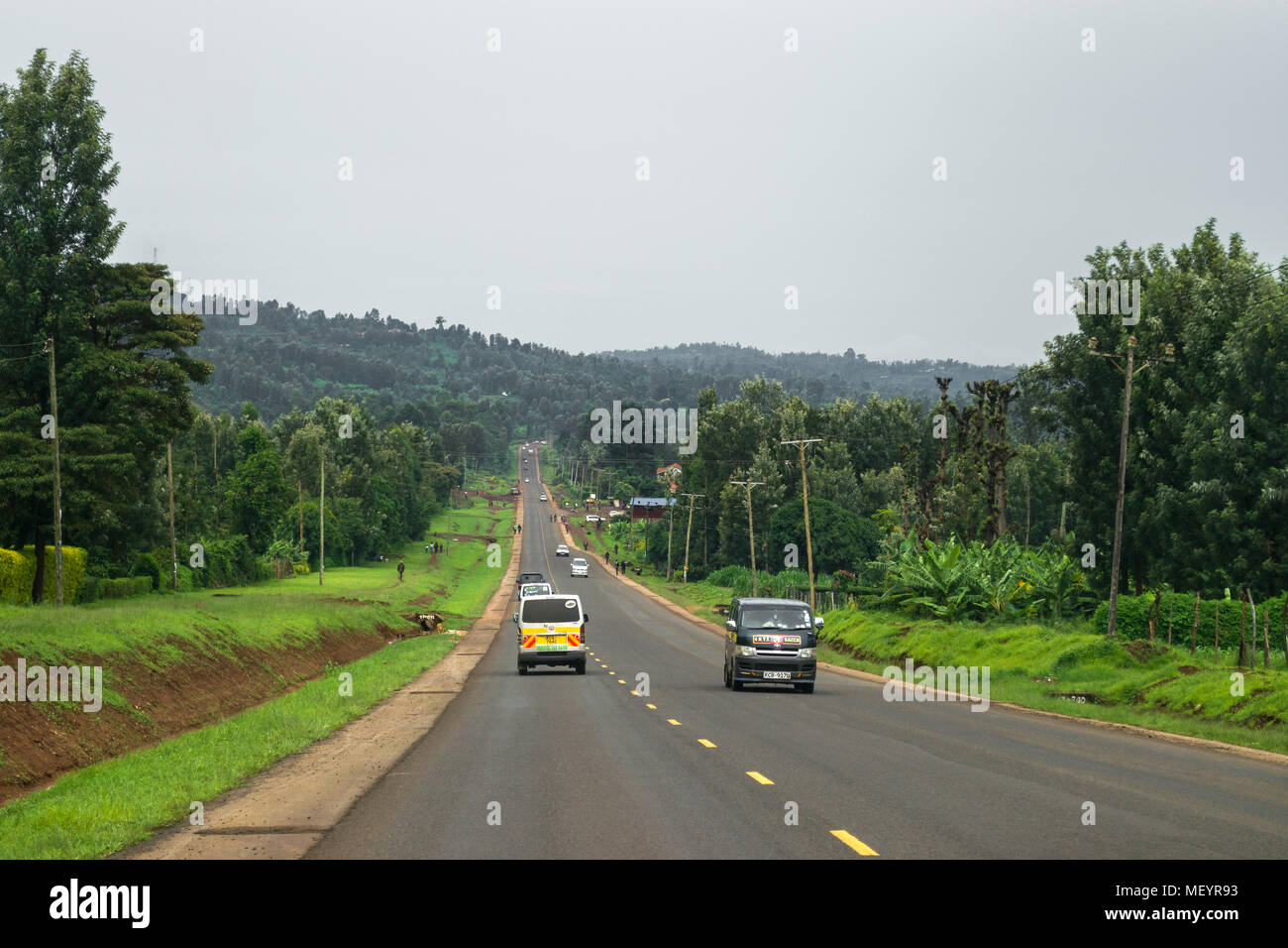 A typical highway with vehicles and people on it, lined by trees and forest on an overcast day, Kenya Stock Photo