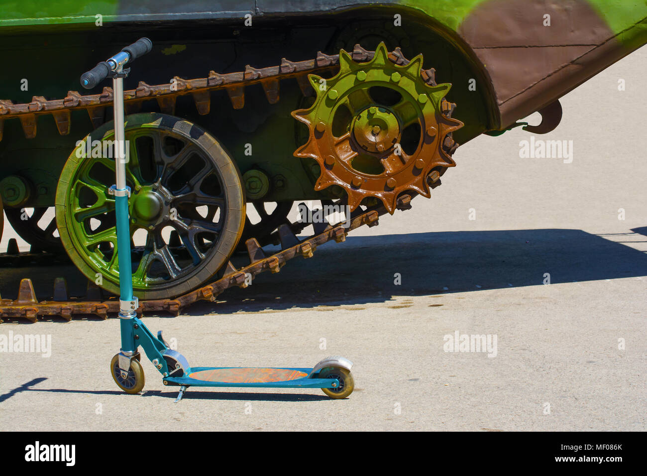 Kids’ scooter in front of a part of a caterpillar track on a military amphibious vehicle. The vehicle has a verdant camouflage scheme on it. Stock Photo