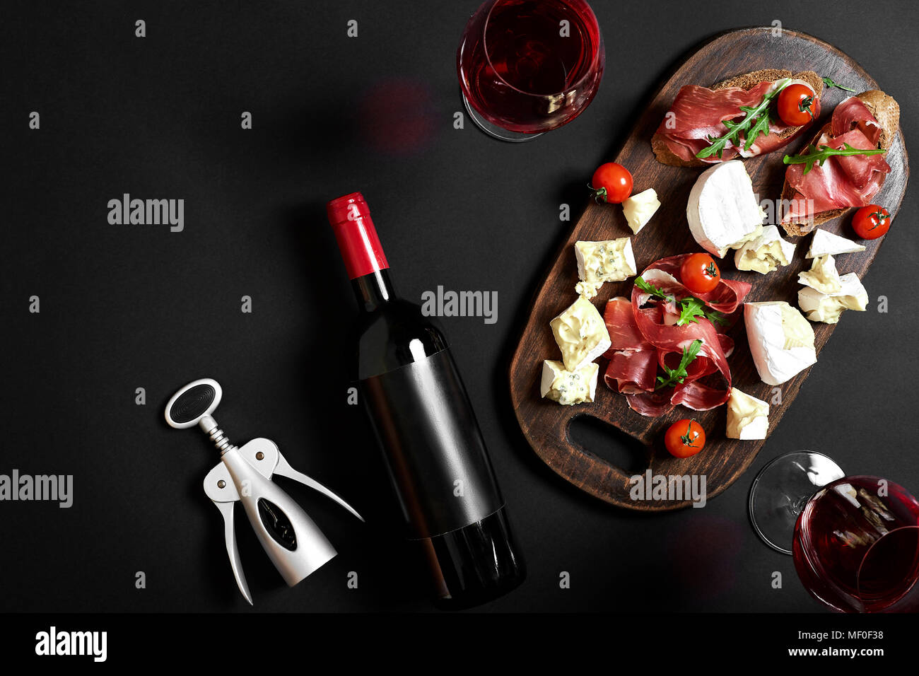 Delicious appetizer to wine - ham, cheese, baguette slices, tomatoes, served on a wooden board, and glass with red wine on black surface Stock Photo
