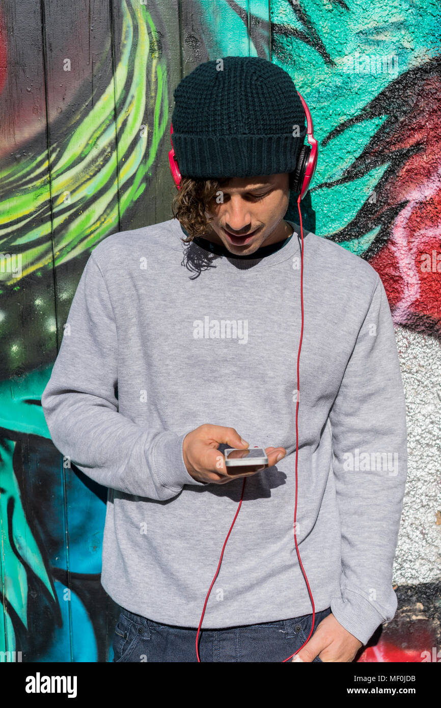 Young man in front of graffity wall hearing music with headphones while using cell phone Stock Photo