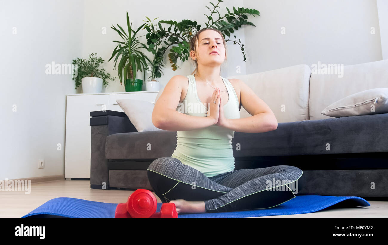 Beautiful young woman sitting on fitness mat at living room and ...