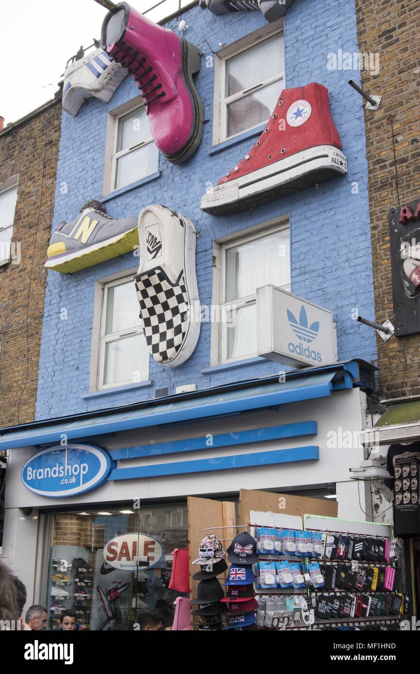 Low angle photograph showing three dimensional, giant shoes, including  Nike, New Balance, Van's, All Star's, Dr Martens, and Adidas, decorating  the brick facade of the 'Brandshop.co.uk' storefront, on a street located at