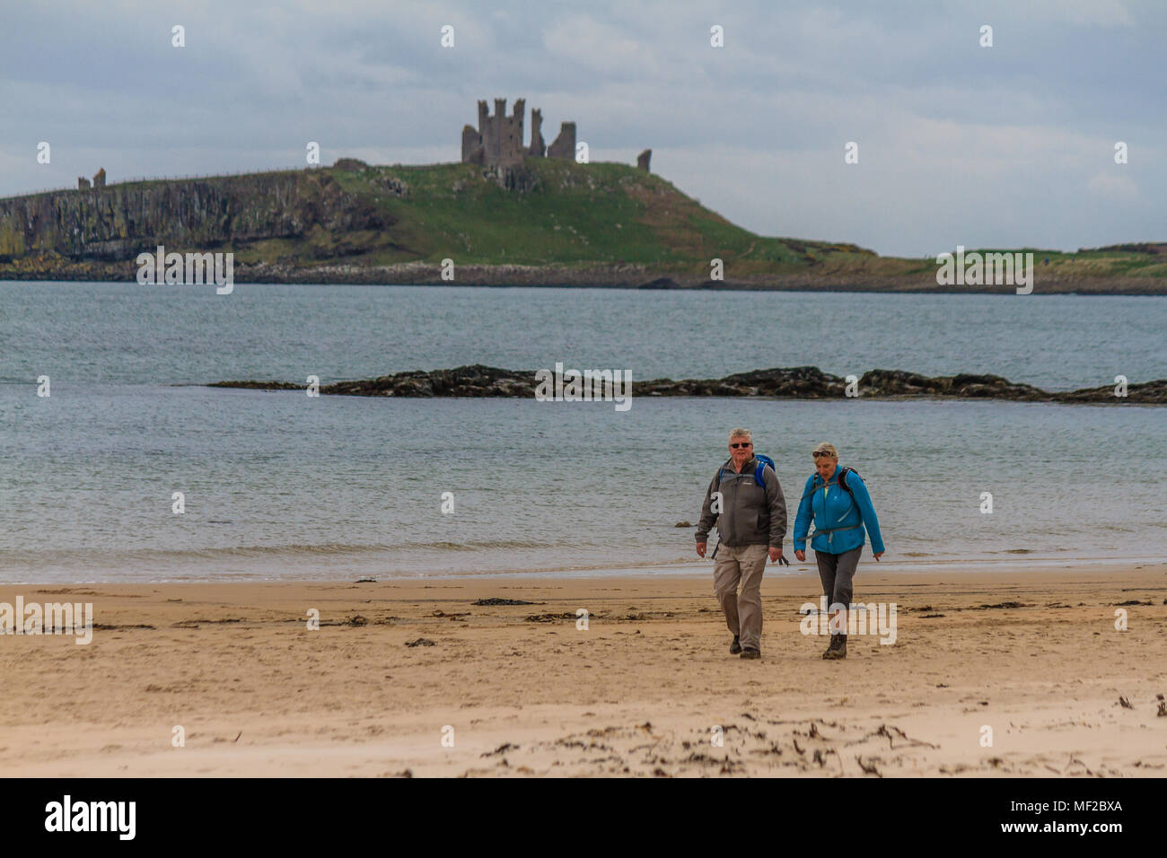 A couple walk along Embleton Beach past Dunstanburgh Castle in Northumberland, UK. Stock Photo