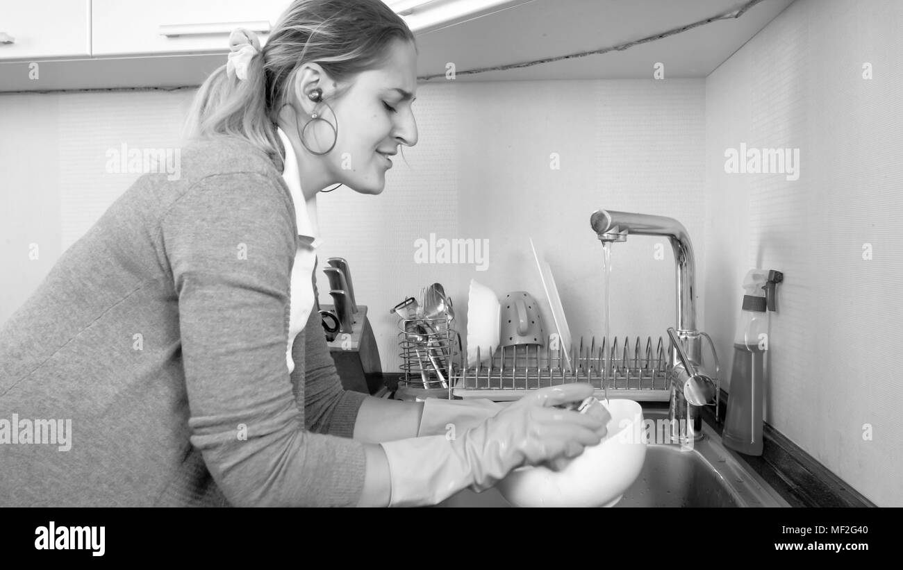 Black and white portrait of smiling young housewife washing dishes and listening music with earphones Stock Photo