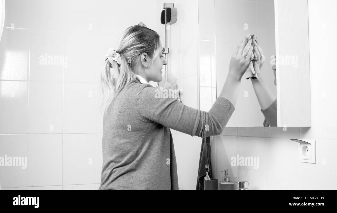Black and white image of young housewife cleaning stains from bathroom mirror Stock Photo