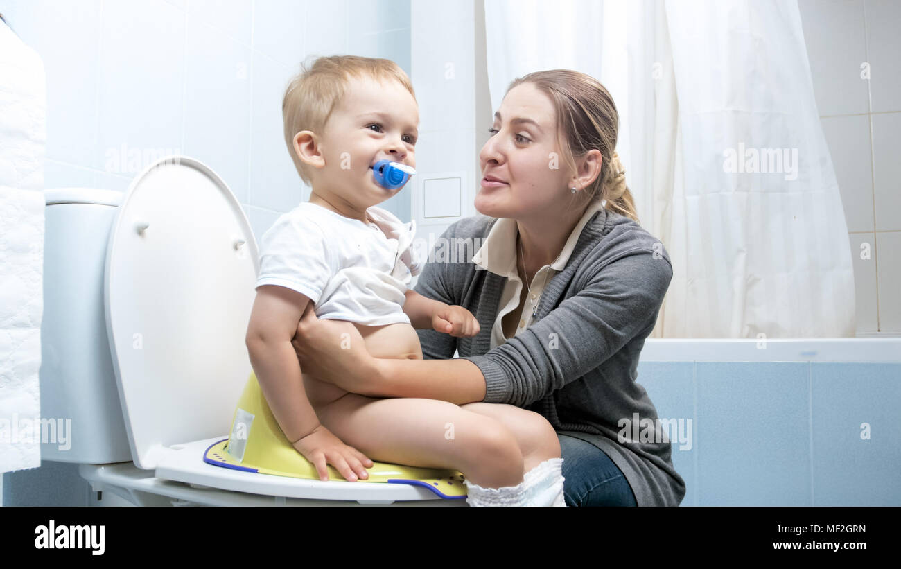 Cute smiling toddler boy sitting on toilet with young mother Stock Photo