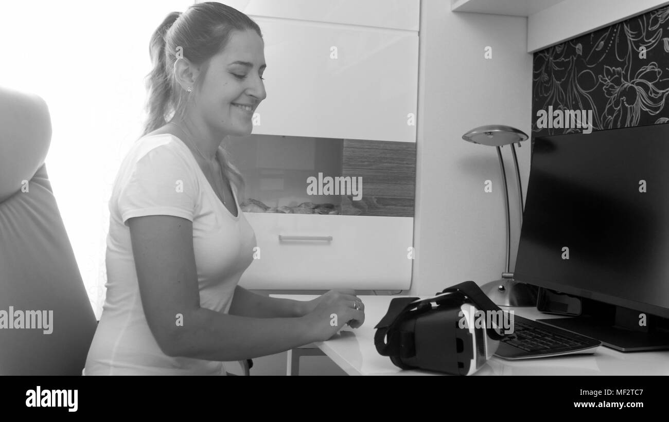 Closeup black and white portrait of smiling young woman looking at vr headset lying on table Stock Photo