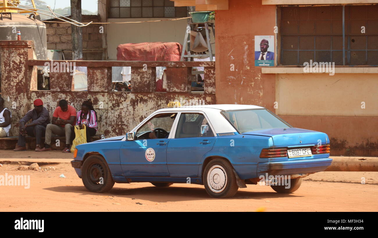 Blue taxi driving in Bissau, Guinea Bissau and people sitting on the side of the road. Stock Photo