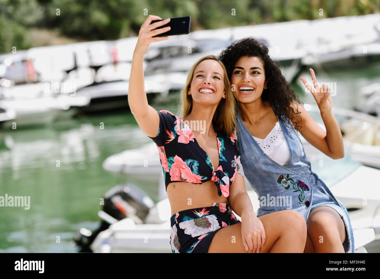 Spain, Andalusia, Marbella. Two multiracial traveler women taking a selfie photograph with smart phone together in boat harbor. Lifestyle concept. Stock Photo