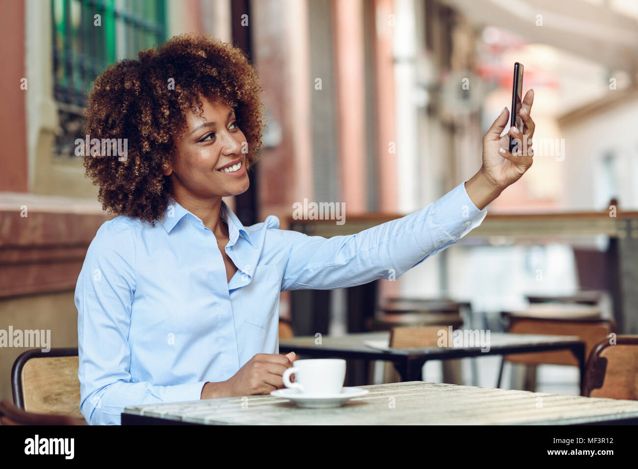 Spain, Andalusia, Malaga.Black woman, afro hairstyle, taking a selfie photograph with a smartphone, sitting in a coffee shop. Happy girl smiling in ur Stock Photo
