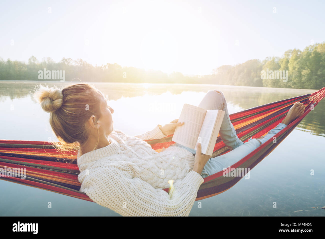 Young woman relaxing on hammock by the lake at sunrise reading a book. People travel relaxation wellbeing learning concept. France, Europe Stock Photo