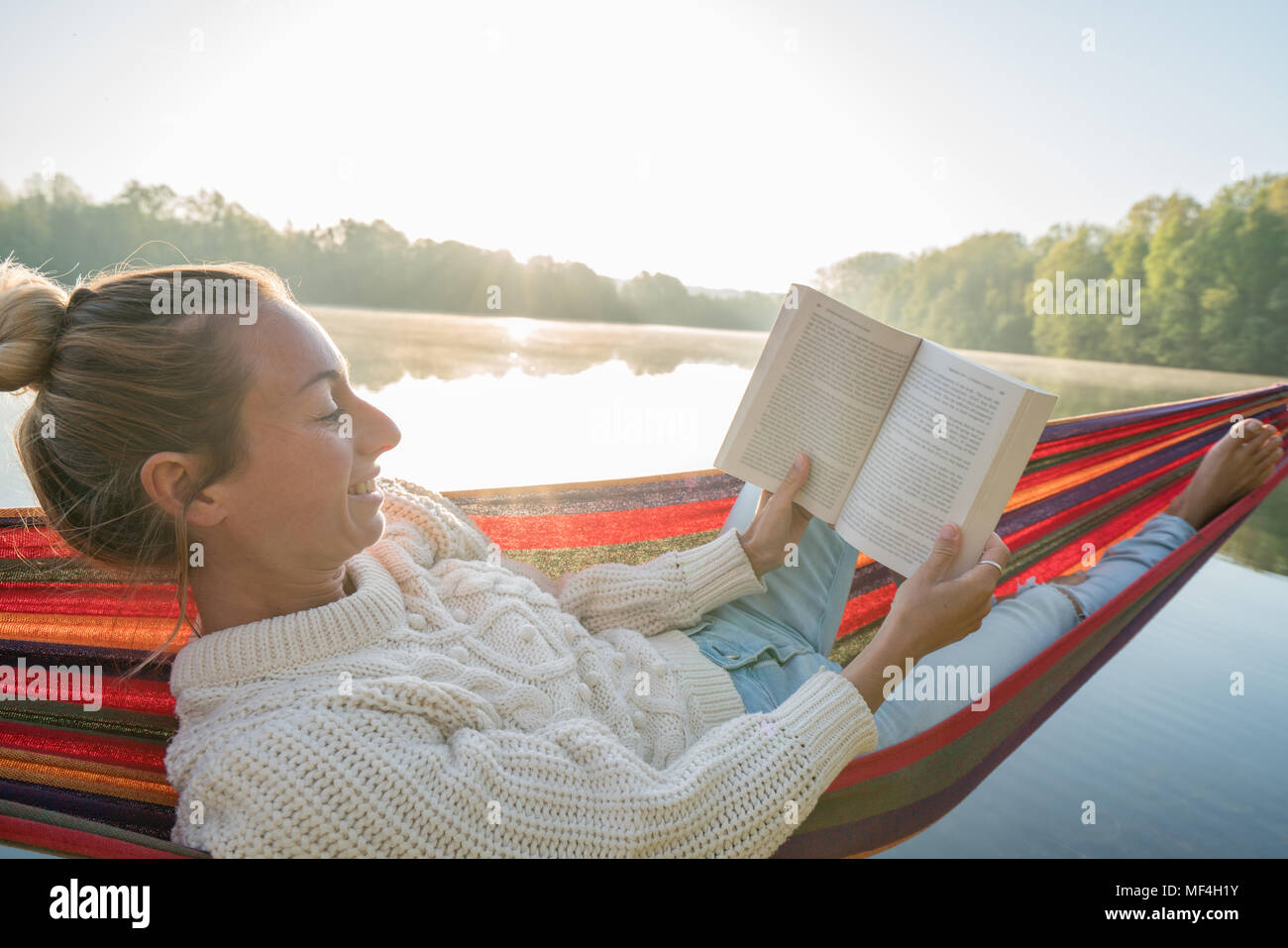 Young woman relaxing on hammock by the lake at sunrise reading a book ...