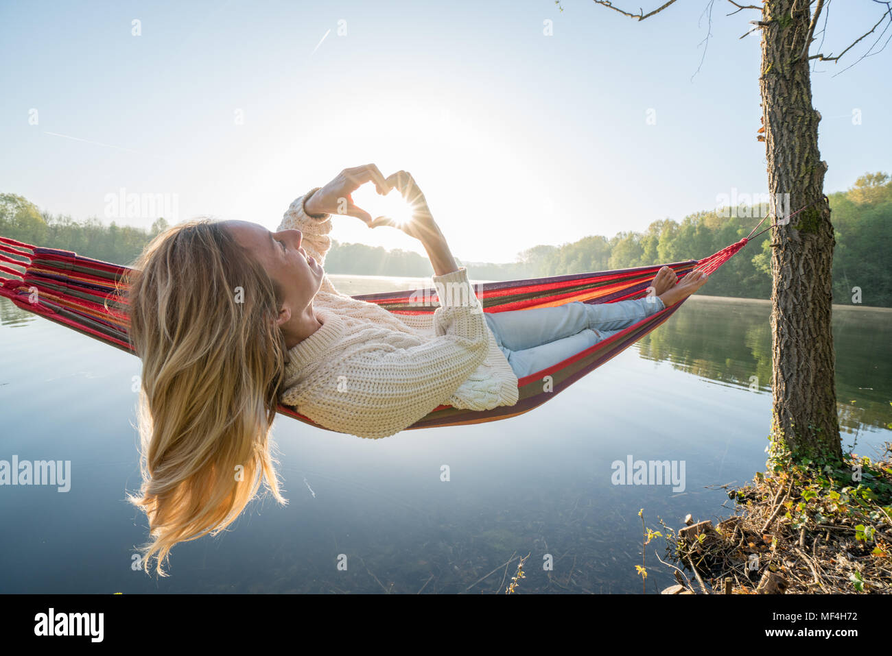 Young woman relaxing on hammock making a heart shape finger frame on beautiful lake landscape, love people romance concept. France, Europe Stock Photo