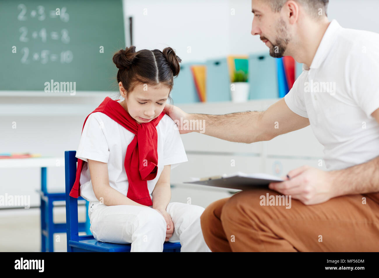 Pretty little girl sitting in classroom and crying because of bad mark while professional male teacher comforting her Stock Photo