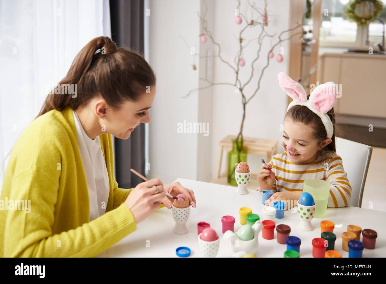 Happy mother and daughter with bunny ears sitting at table painting Easter eggs Stock Photo