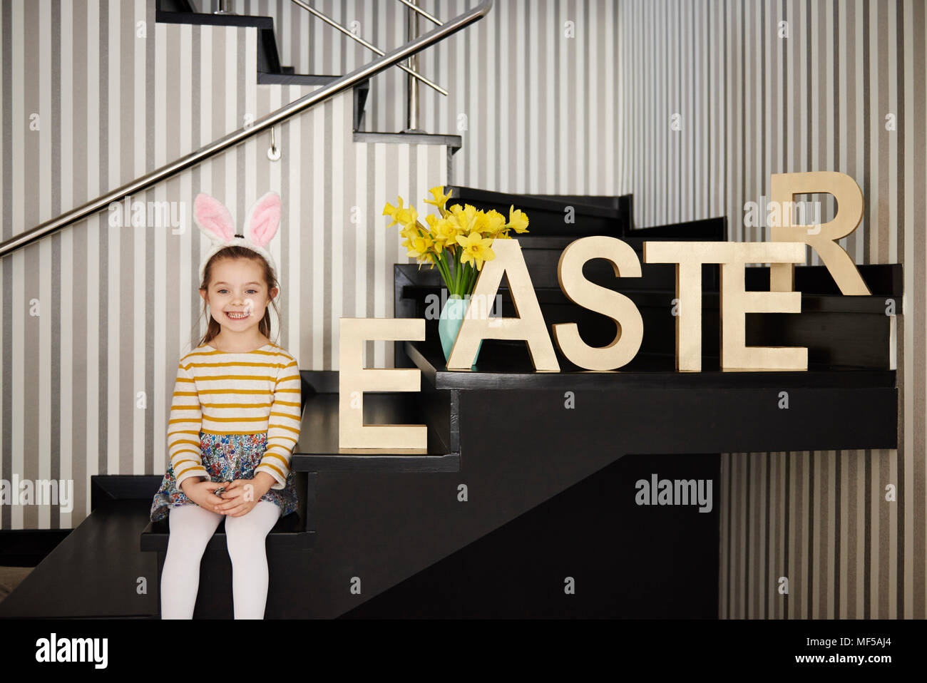 Portrait of smiling girl with bunny ears sitting on stairs next to word 'Easter' Stock Photo