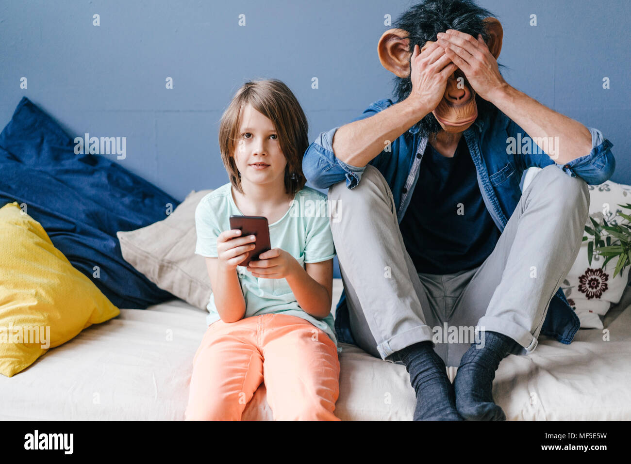 Father wearing monkey mask sitting next to son using smartphone at home Stock Photo