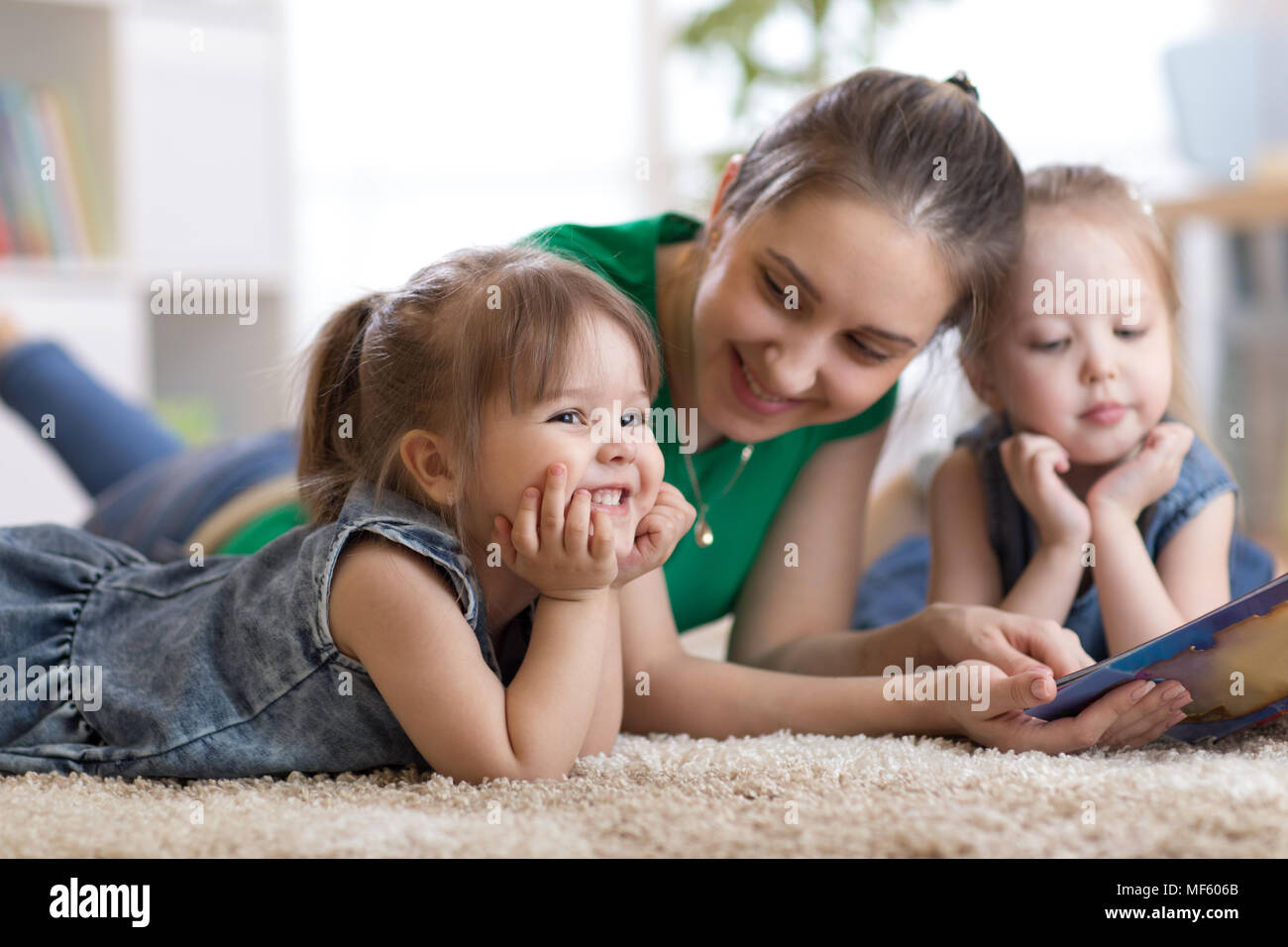 Children laughing and having fun reading stories with their mother laying on the floor at home Stock Photo