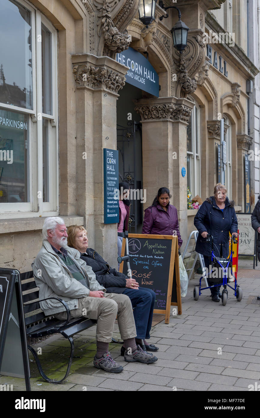 A couple relaxing by sitting on a bench outside The Corn Hall in Cirencester with shoppers exiting the arcade Stock Photo