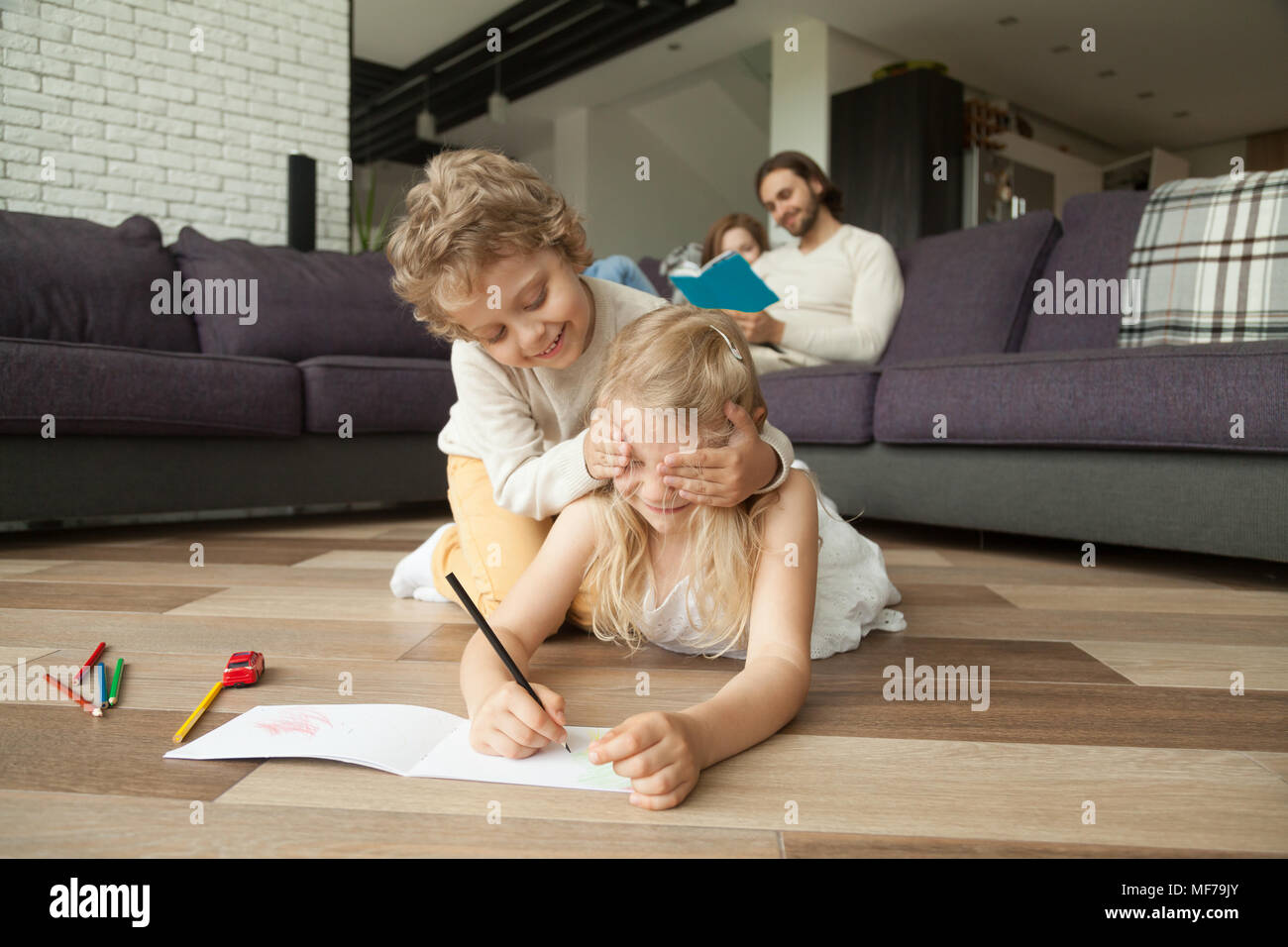 Siblings playing on floor while parents reading book on sofa Stock Photo