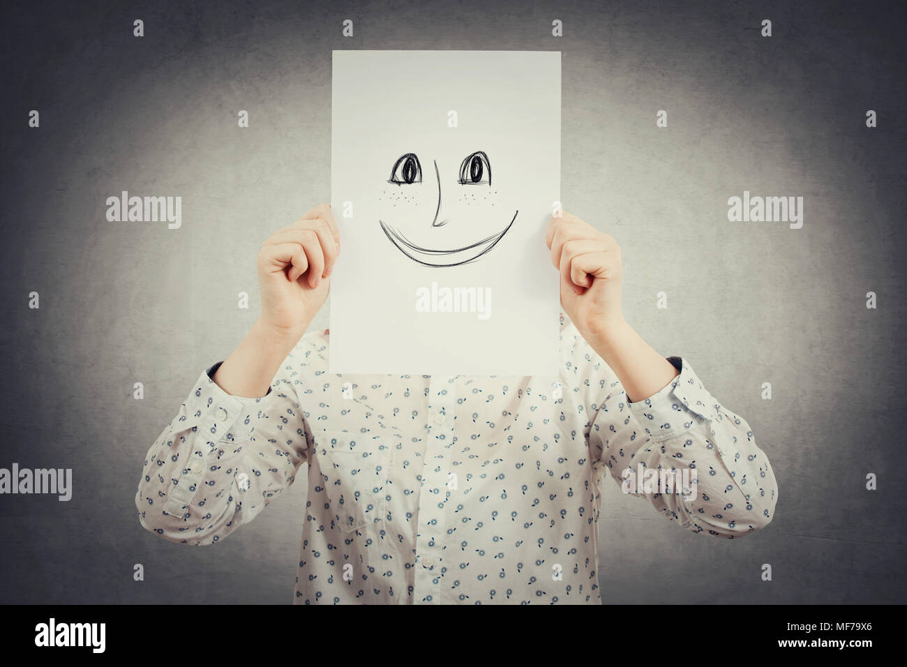 Young businesswoman covering her face using a sheet with drawn happy, smiling emoticon sketch, like a mask to hide her real emotion from society. Crea Stock Photo