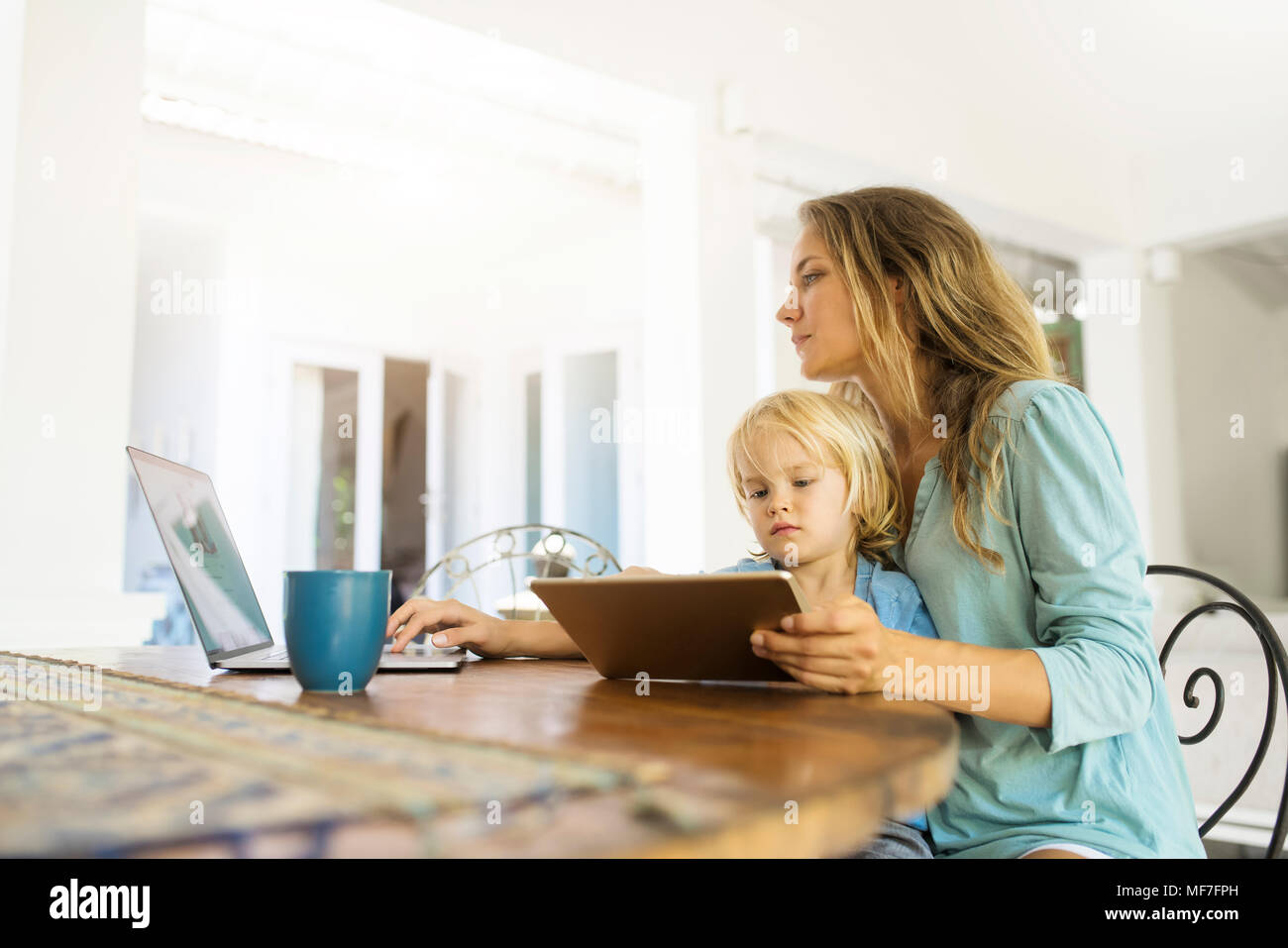 Boy sitting on his mother's lap and looking at a tablet while his mother is working on a laptop Stock Photo