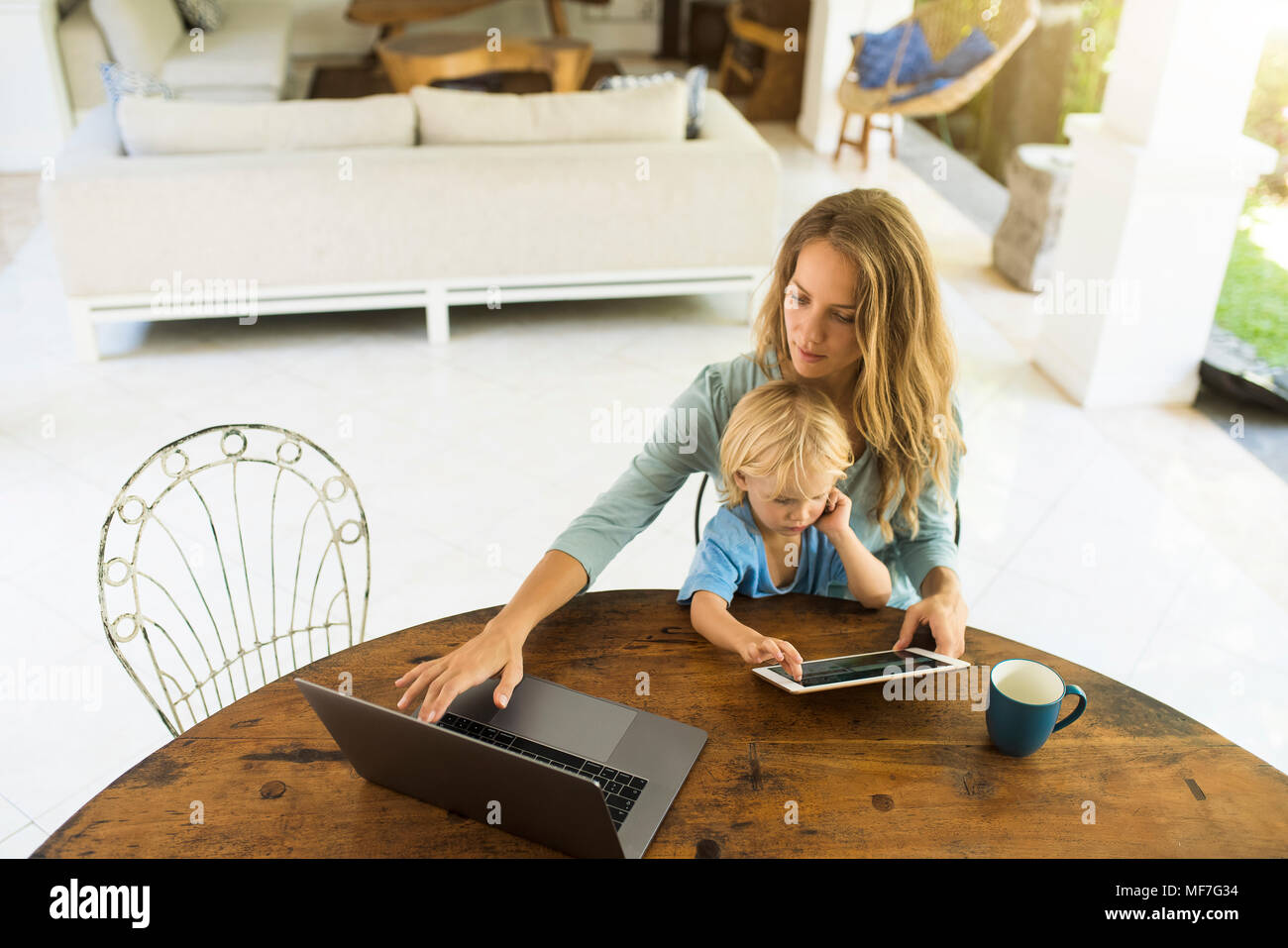 Boy sitting on his mother's lap and looking at a tablet while his mother is working on a laptop Stock Photo