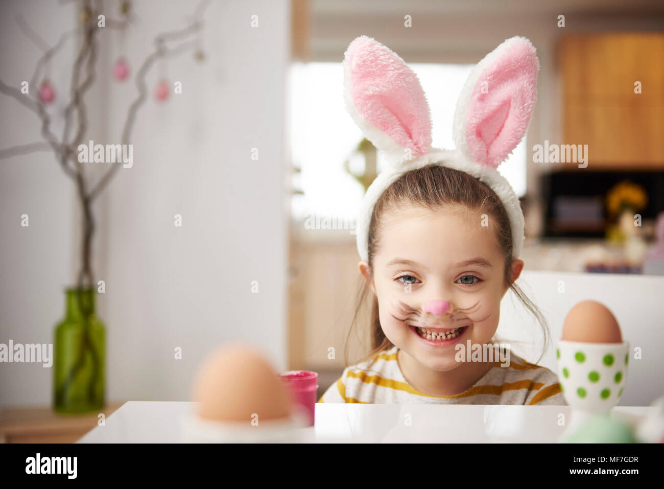 Portrait of smiling girl with bunny ears sitting at table with Easter eggs Stock Photo