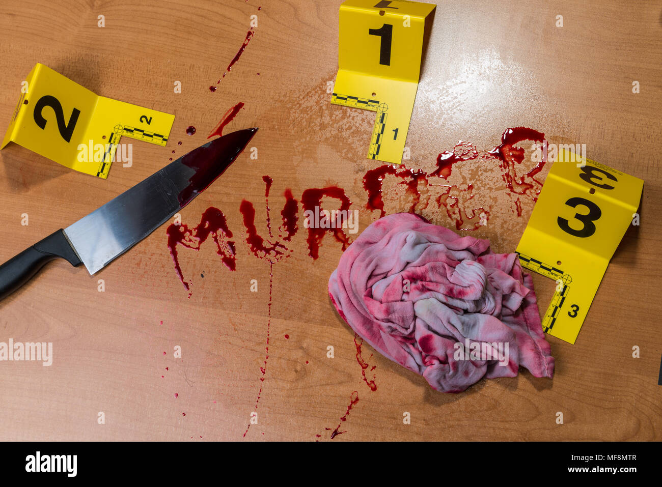 The word “murder” written in blood on a wood surface beside a bloody knife and a bloody rag, all marked by crime scene evidence markers. Stock Photo