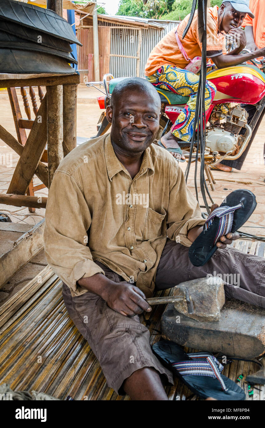 Smiling African man sitting on ground and manufacturing tire flip flops in street Stock Photo