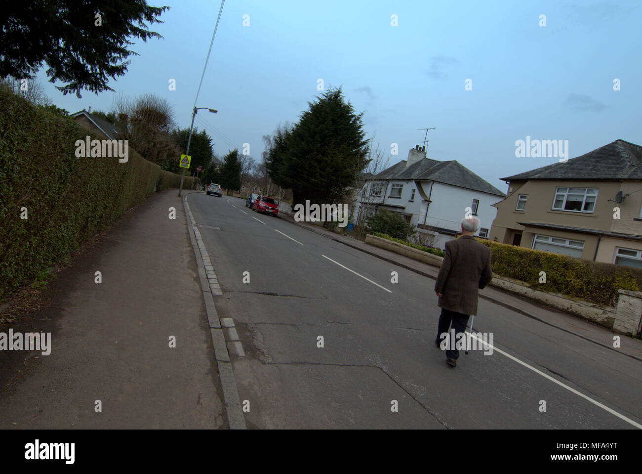 wide angle street view of old man crossing middle of  road with walking stick urban middle class housing Stock Photo