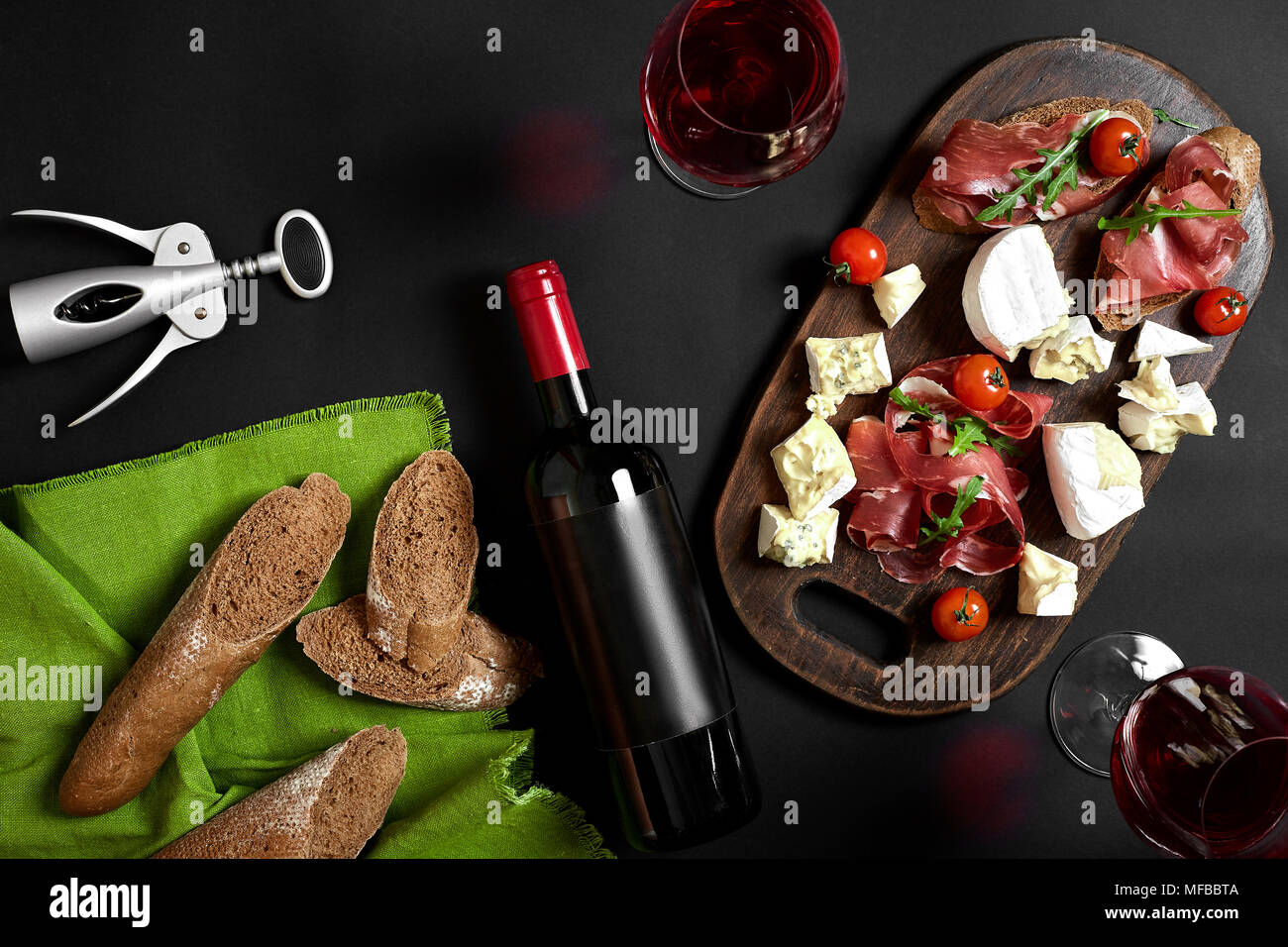 Delicious appetizer to wine - ham, cheese, baguette slices, tomatoes, served on a wooden board, and glass with red wine on black surface Stock Photo