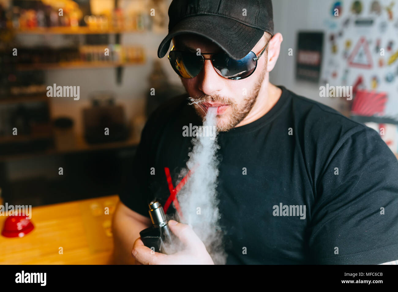 Brutal bearded man in sunglasses smoke an electronic cigarette and releases clouds of vapor at the vape shop. Stock Photo