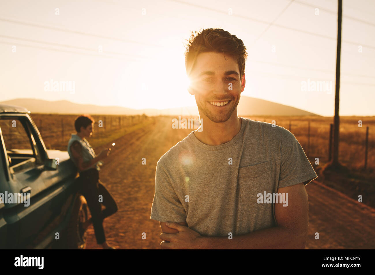 Close up of smiling man standing on a mud track with his partner in the background. Couple enjoying their road trip in country side Stock Photo