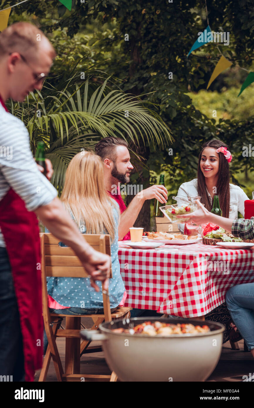 A man grilling food in front of his friends sitting by a table with red and white checkered tablecloth at BBQ party in the garden Stock Photo