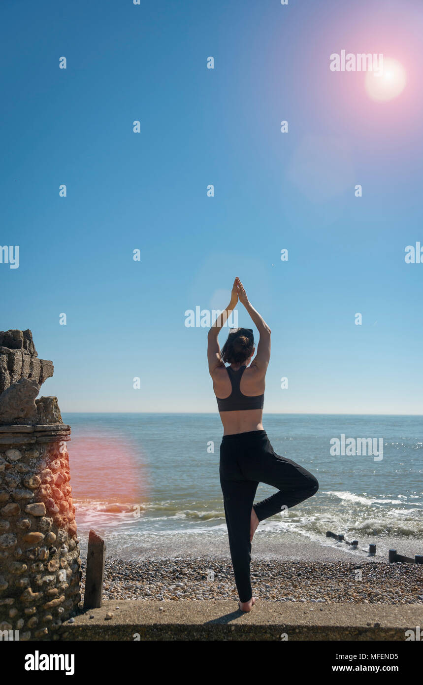 woman practicing yoga by the sea in the sunshine, lens flare. Stock Photo