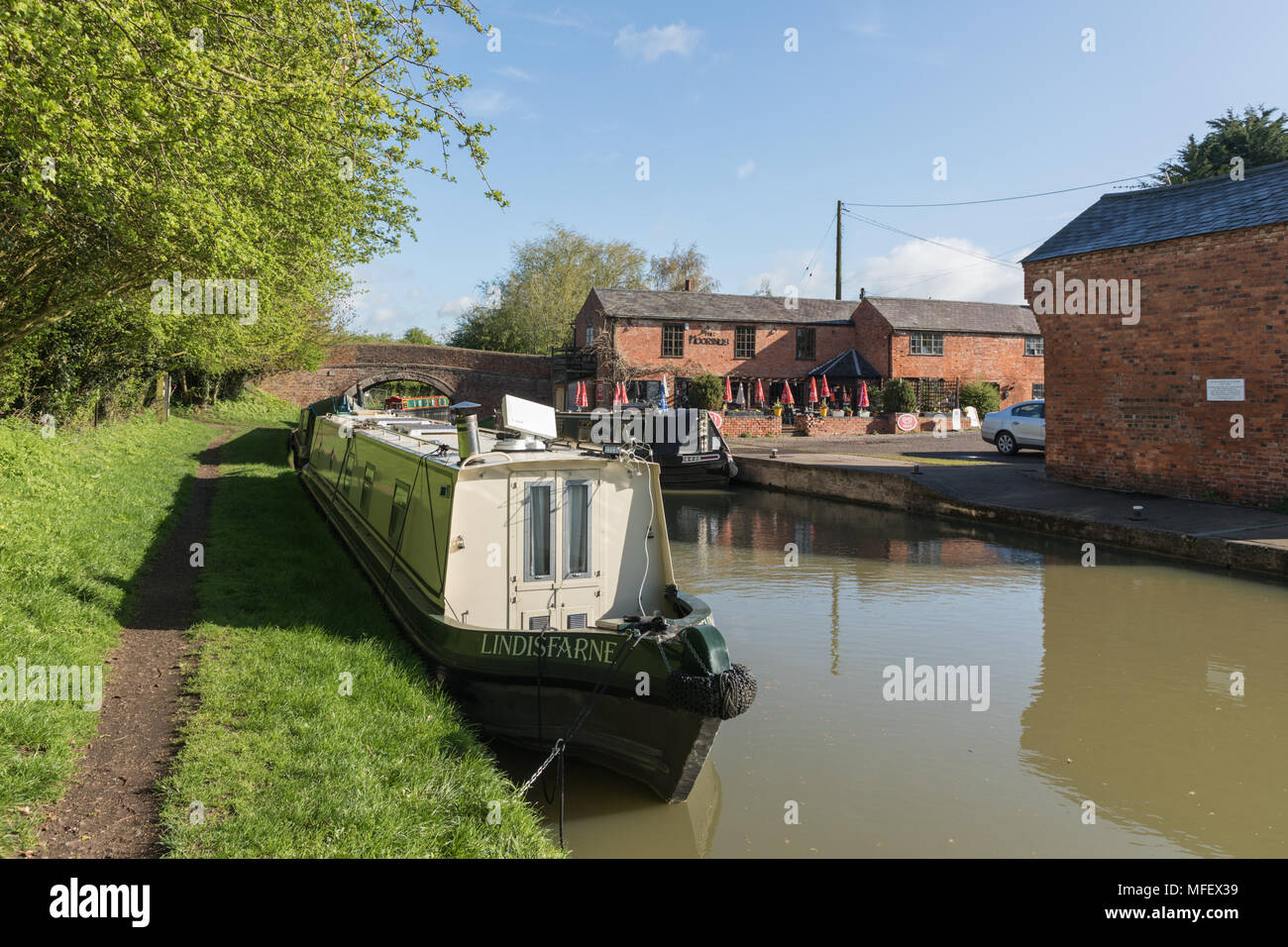 Narrowboats, lit by the spring morning sunshine, are moored at Crick Wharf. Furled umbrellas are in the garden of the restaurant on the opposite bank. Stock Photo