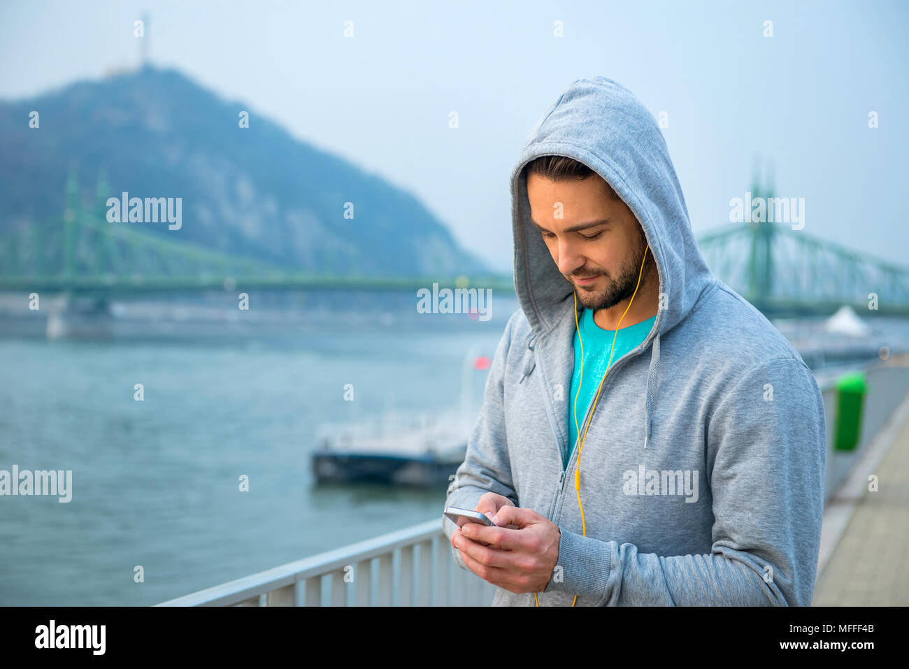 A handsome young man standing in training clothes and using his phone with earphones at the riverside Stock Photo