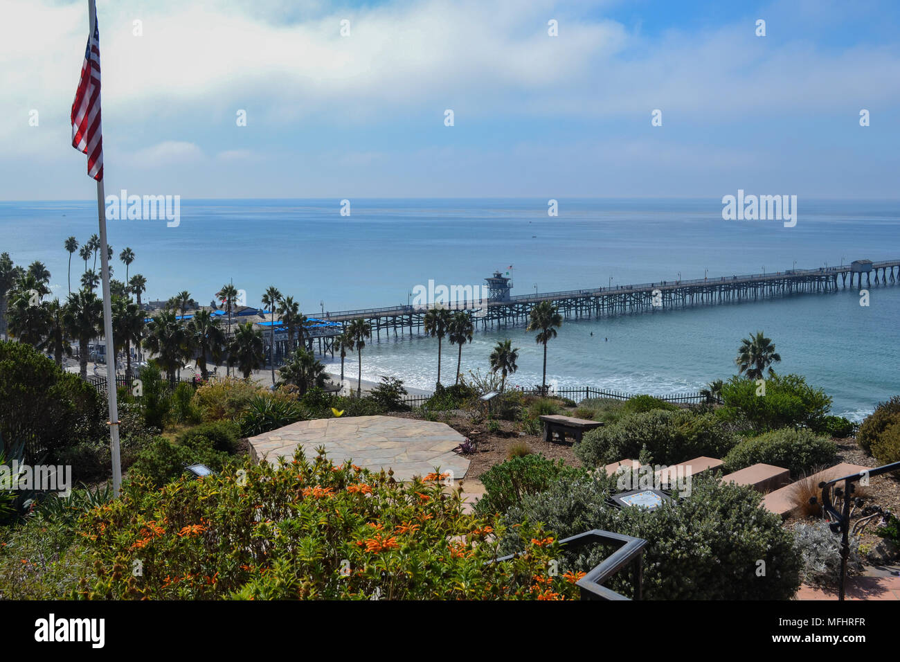 San Clemente Pier, Southern California, on a clear day, American US Flag on flagpole in the foreground, blue sky with clouds, Pacific Ocean shoreline Stock Photo