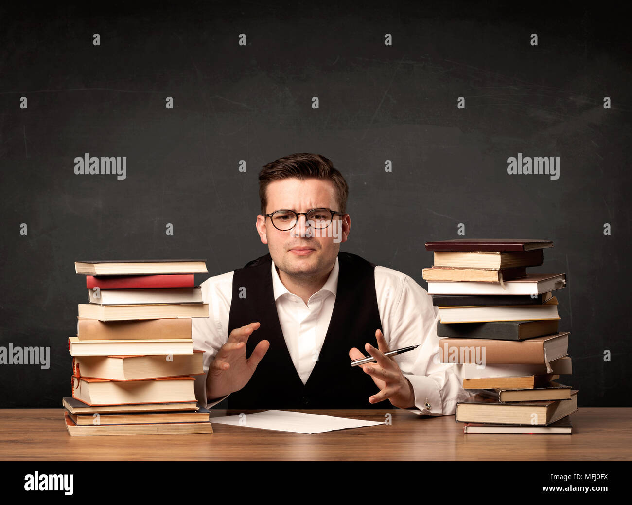 A young teacher in glasses sitting at classroom desk with pile of books ...