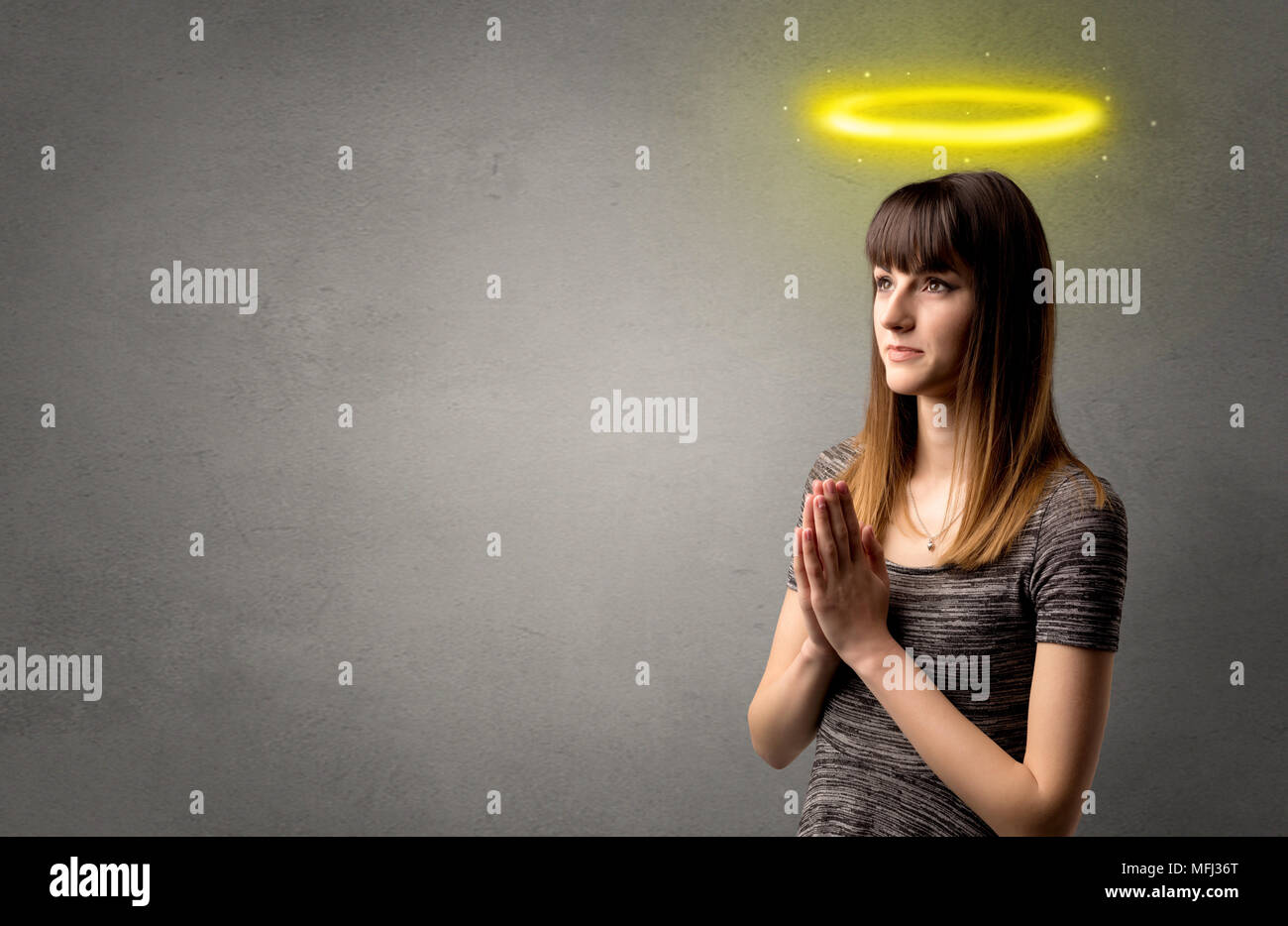 Young woman praying on a grey background with a shiny yellow halo above ...